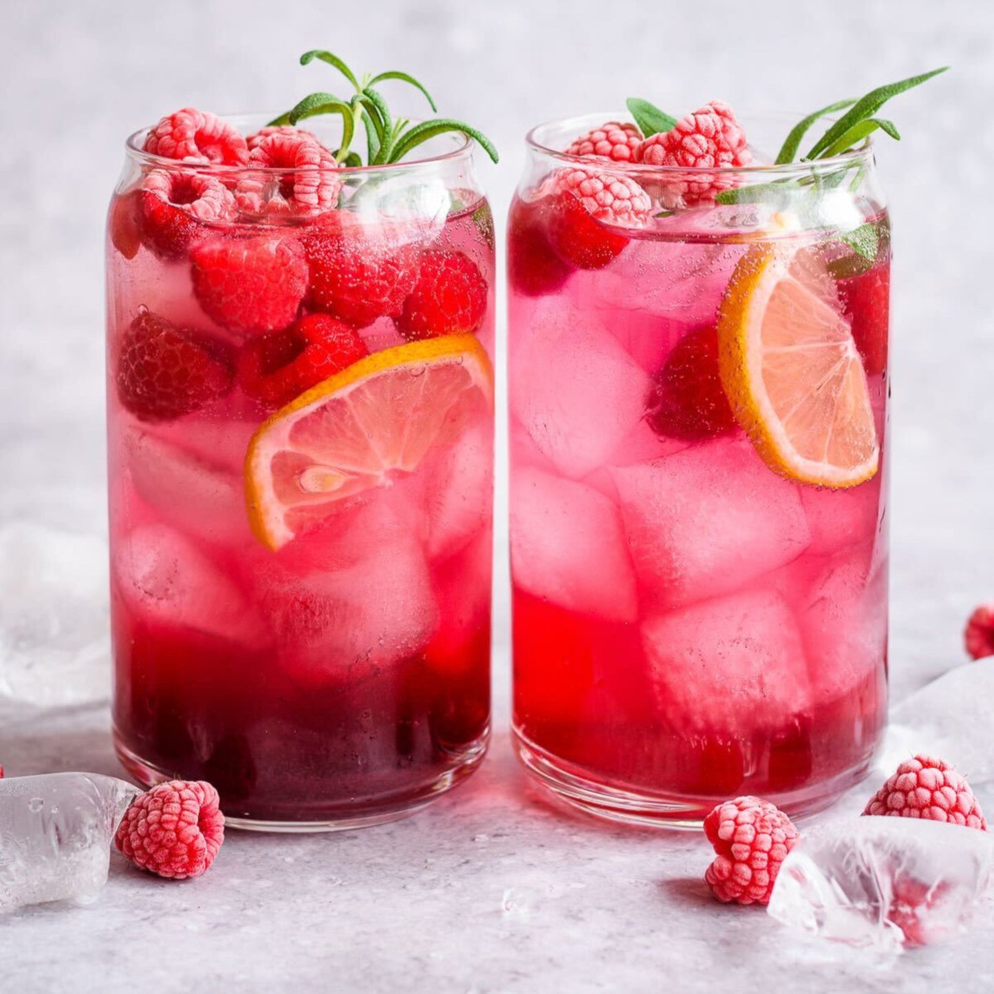 Two glasses of raspberry lemonade with ice and garnishes on a light background