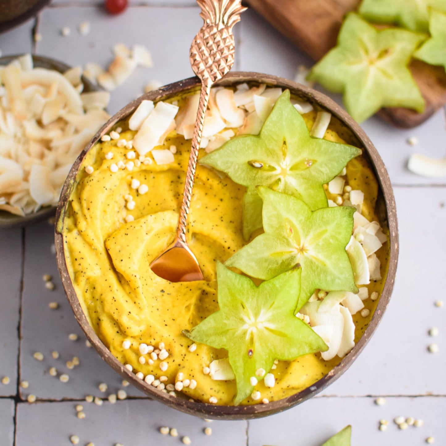 Coconut bowl with yellow smoothie, star-shaped fruit, and a spoon on a light wooden surface.